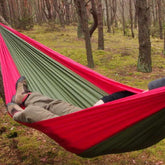 Person relaxing in a Bushmen ZEN Hammock in a forest, showcasing its red and green parachute nylon design.